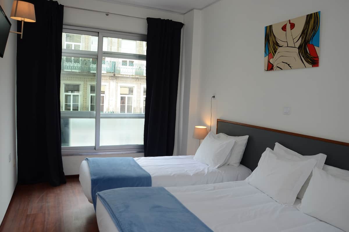 A hotel room with two beds, a window with curtains, a wall-mounted lamp, and a painting.