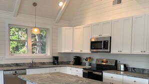 A kitchen with white cabinets, a granite countertop, a central island with stools, a window, and an air conditioning unit. - New Listing - Island View Cottage (Phippsburg)