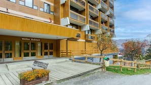 A modern building with a wooden facade, a wooden deck with a bench and flower bed, and a sign reading "Vostok Zodiac." - Vostok Zodiaque by Interhome (Le Corbier)