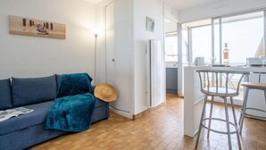 A modern apartment with a blue sofa, a hat on the floor, a kitchen area with bar stools, and a large window. - Apartment Milos by Interhome (Canet-Plage)