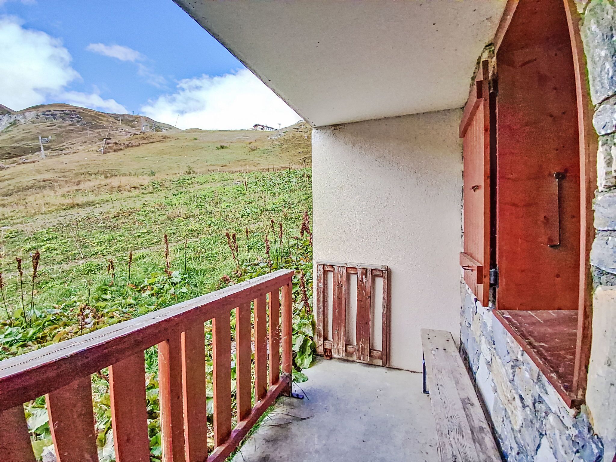 A balcony with a stone wall, wooden railing, and a view of a grassy field and hills.