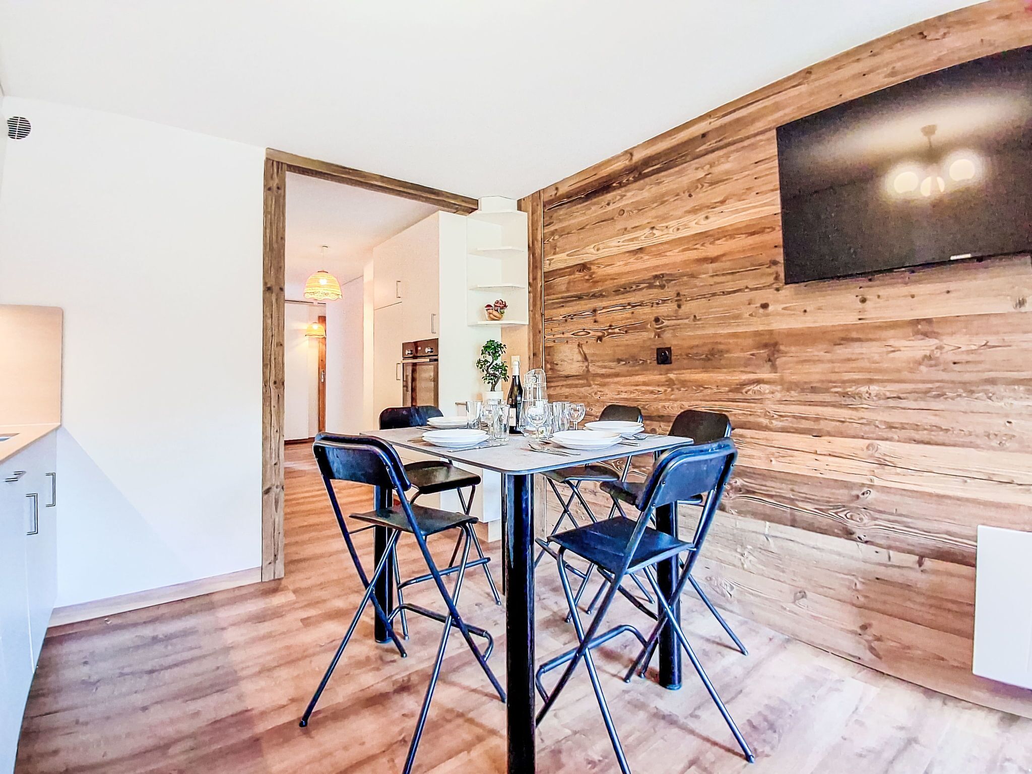 A dining area with a wooden wall, a TV mounted above the table, and a door leading to another room.