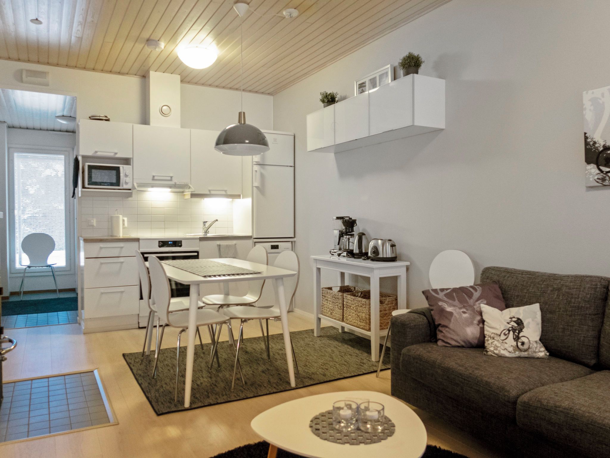 Un ambiente compatto con angolo cottura, zona pranzo con tavolo e sedie, divano con cuscini decorativi e un piccolo tavolino con bollitore.