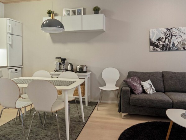 Una sala de estar moderna con un sofá gris, sillas blancas, una mesa de comedor y una zona de cocina con electrodomésticos.
