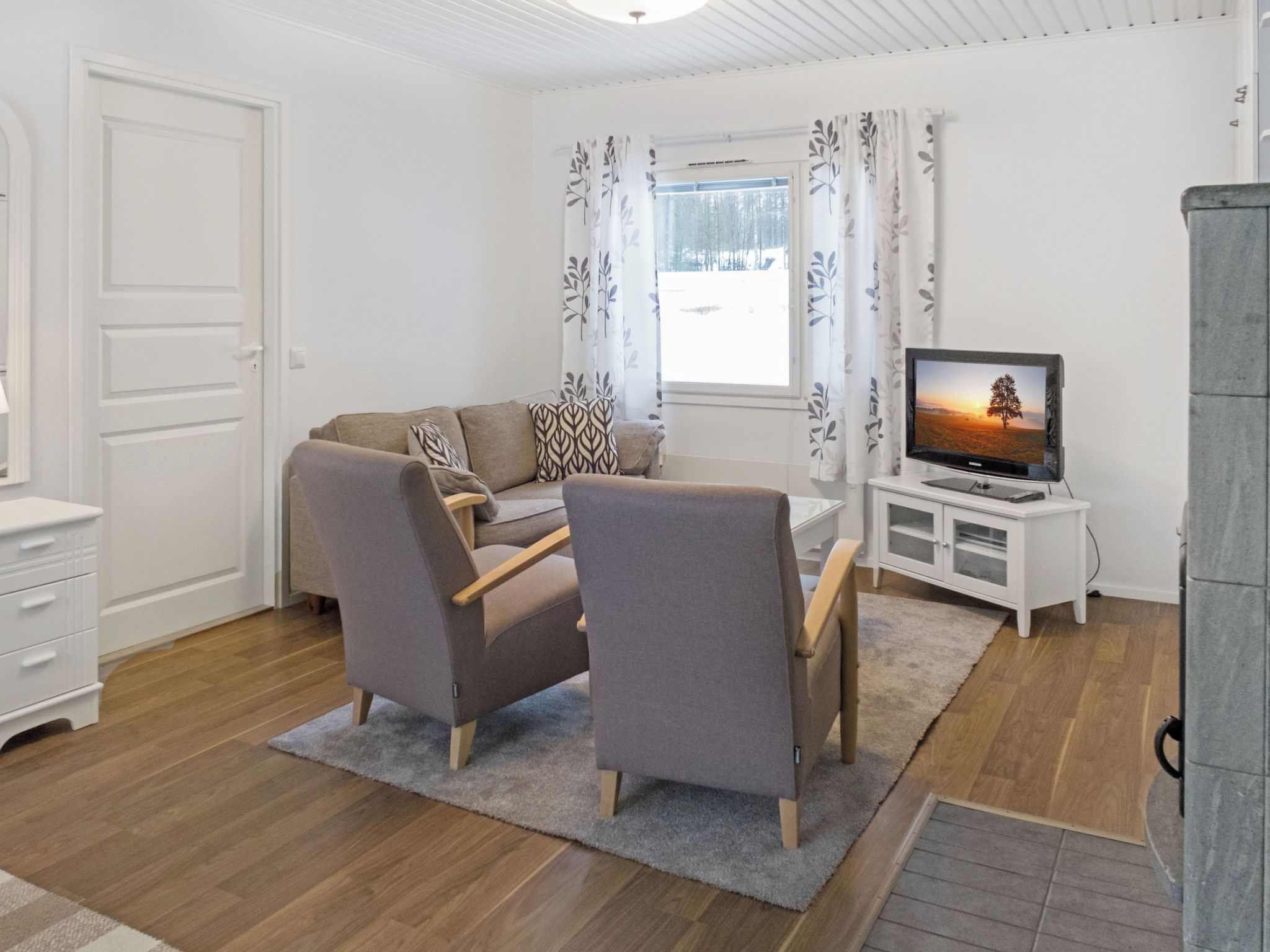 A living room with a sofa, two armchairs, a TV on a stand, a white cabinet, and a window with curtains.