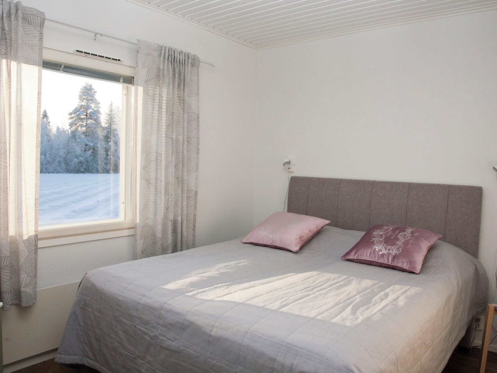 Ein Schlafzimmer mit einem Bett, zwei Kissen, einem Fenster mit Vorhängen und Blick auf eine verschneite Landschaft.