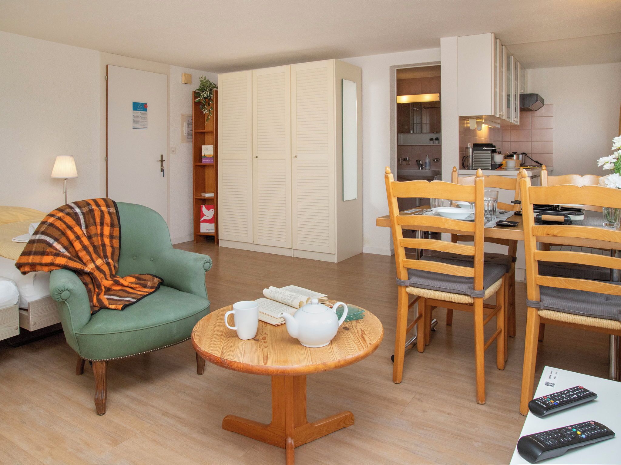 Un salon chaleureux avec un fauteuil vert, une petite table en bois sur laquelle se trouvent une théière et des tasses, un coin repas avec des chaises en bois, et une cuisine visible à travers une porte ouverte.