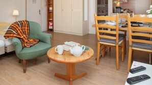 A cozy living room with a green armchair, a small wooden table with a teapot and cups, a dining area with wooden chairs, and a kitchen visible through an open door. - Helene Studio Christel by Interhome (Wengen)