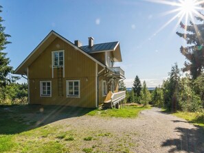 Ein zweistöckiges Holzhaus mit einem Balkon, einer Schotterauffahrt und einer bewaldeten Landschaft.