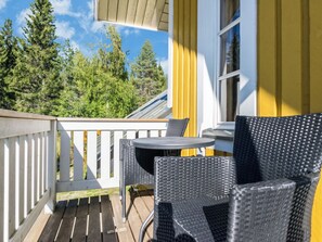 Un balcon doté d’une terrasse en bois, de deux fauteuils en osier, d’une petite table ronde et d’une fenêtre ornée de rideaux jaunes.