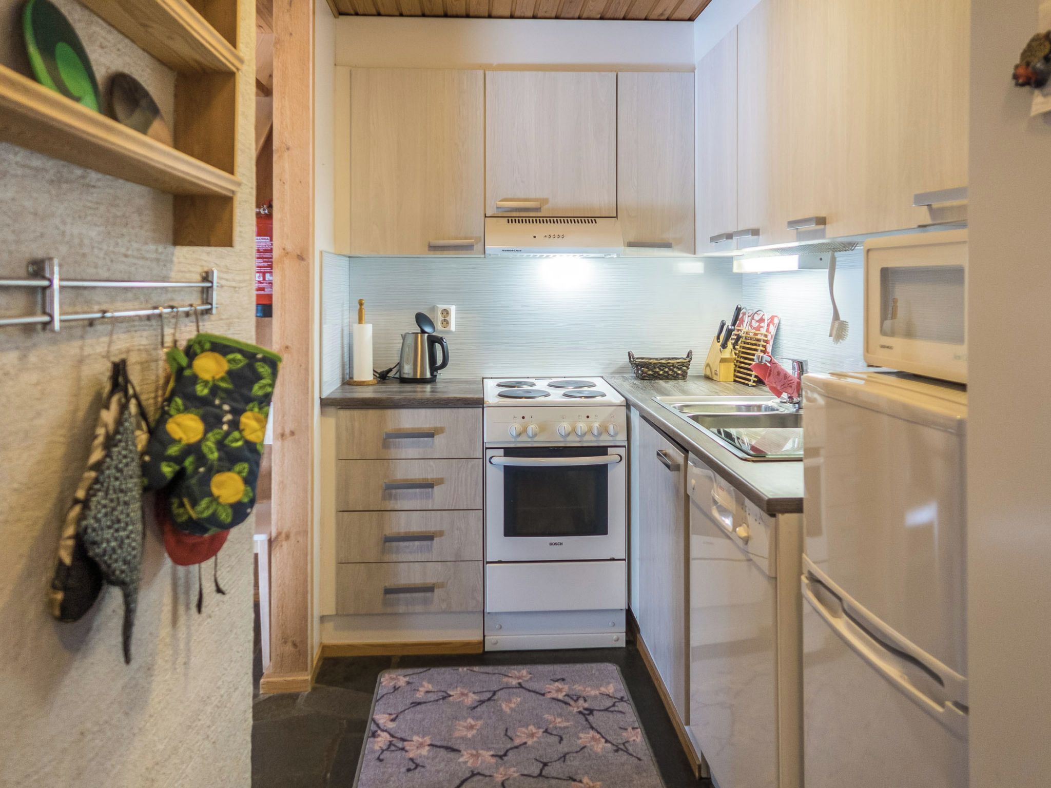 Une cuisine compacte équipée d’une cuisinière Bosch, d’un four à micro-ondes et d’un réfrigérateur. On y trouve des armoires en bois et un pare-feu en carrelage.