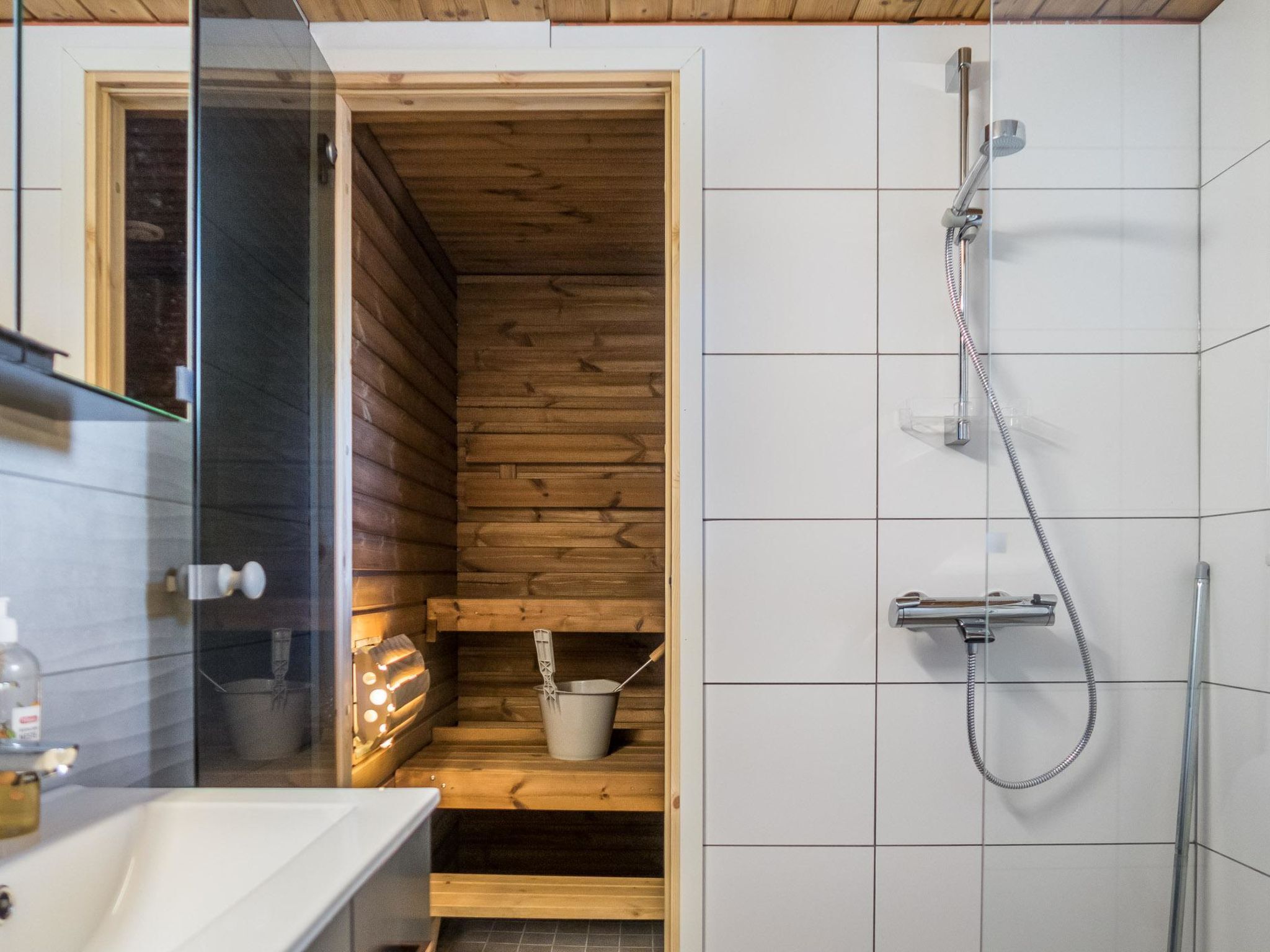 Sauna, jossa on puiset lauteet ja lasiovi, suihkutila letkulla sekä valkoisilla laatoilla päällystetty seinä.