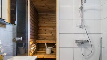 Sauna, jossa on puiset lauteet ja lasiovi, suihkutila letkulla sekä valkoisilla laatoilla päällystetty seinä.