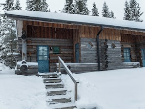 Une cabane en rondins avec un escalier en bois menant à une porte, entourée d’un paysage enneigé et d’arbres.