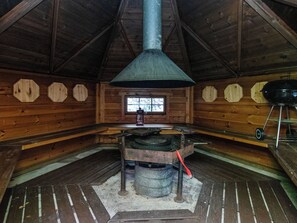 Interior rústico em madeira com uma lareira grande, um fogão a lenha e uma janela com vista para árvores.