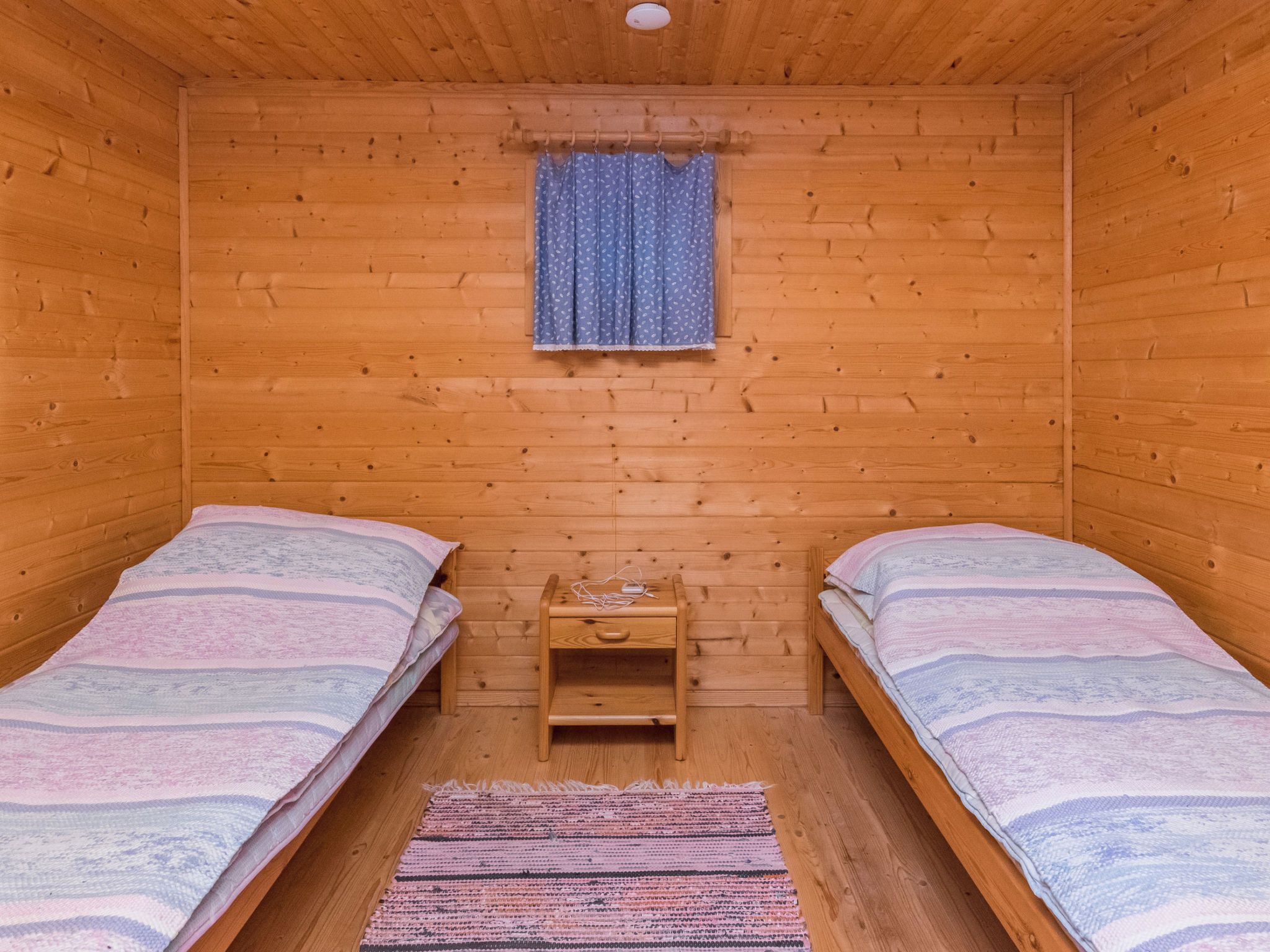 A small wooden room with two single beds, a small wooden nightstand, and a window with a curtain.