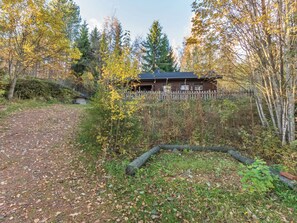 A rustic cabin surrounded by a wooden fence, a dirt path leading to it, and a variety of trees and shrubs in the background.