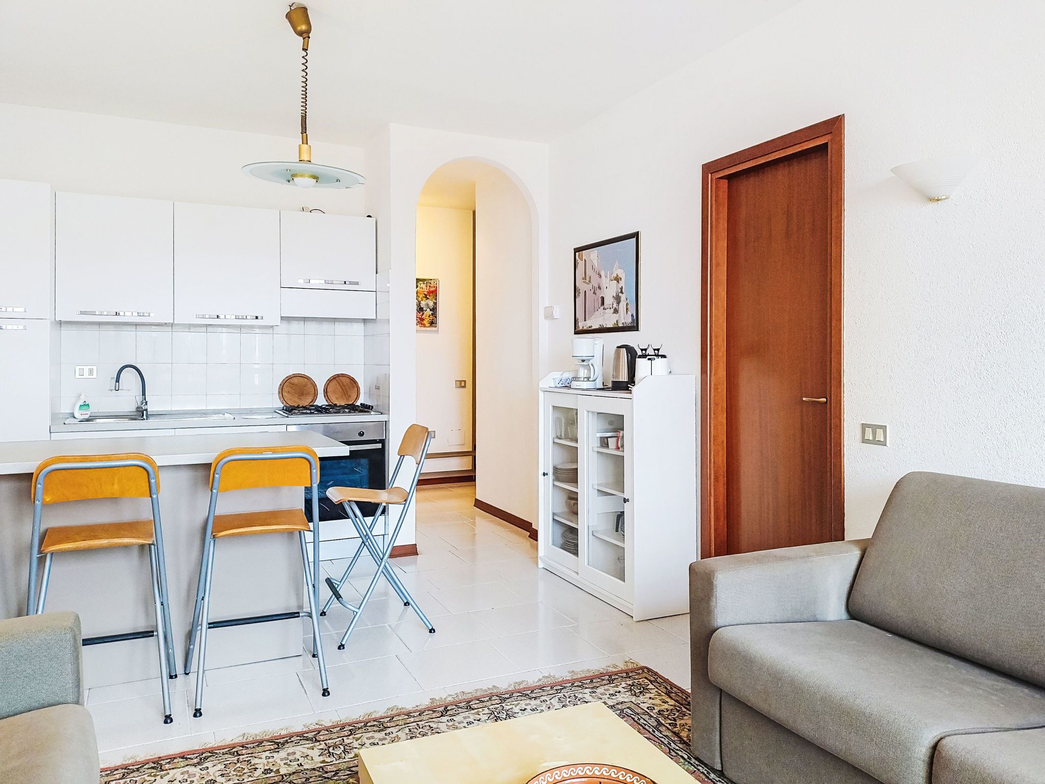 Une cuisine moderne avec des meubles blancs, un coin repas avec des chaises jaunes, un canapé gris et une porte en bois.