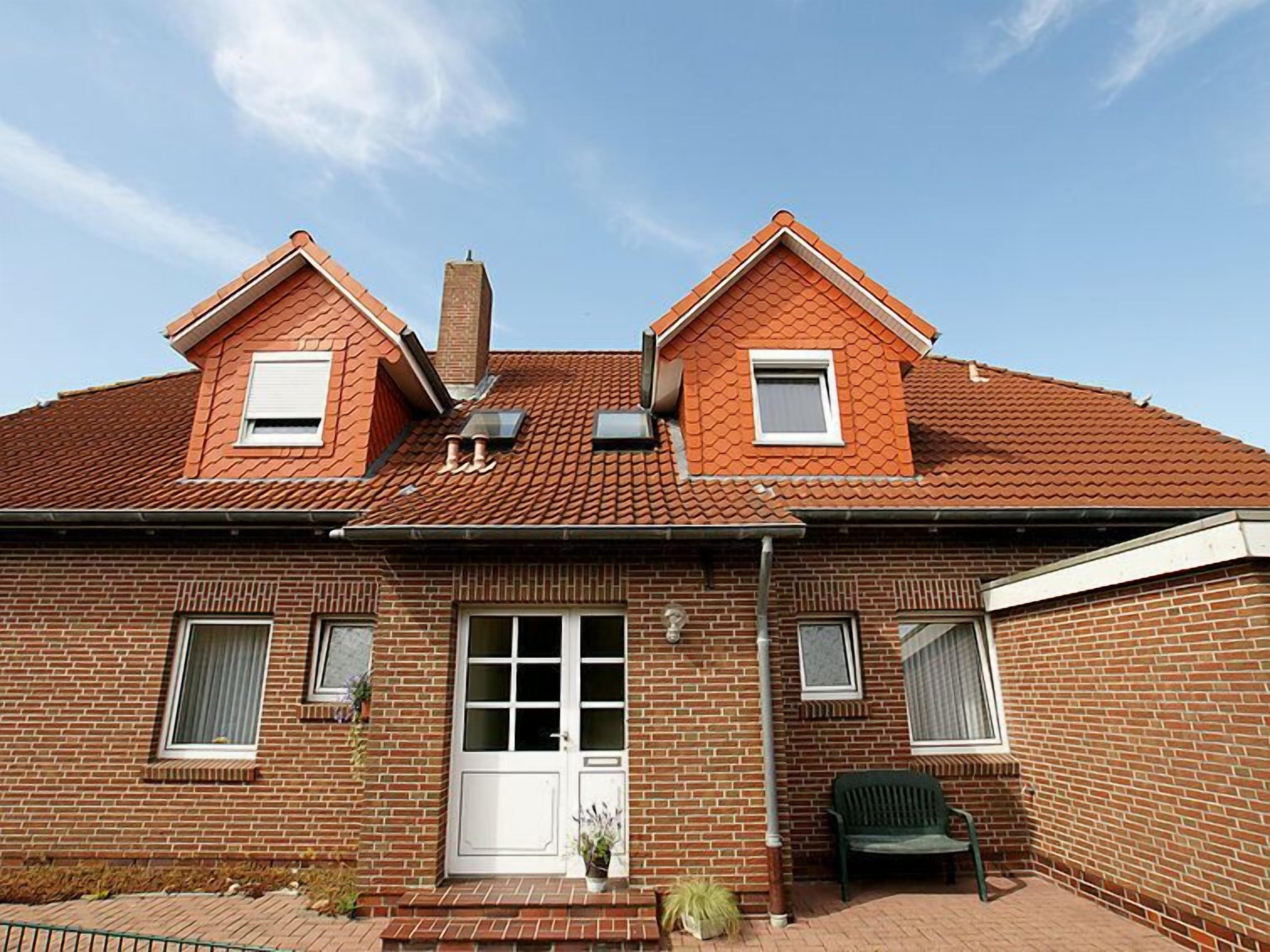 Une maison à deux étages, construite en briques, avec un toit recouvert de tuiles rouges, une entrée centrale dotée d’une porte blanche et deux petites fenêtres à l’étage supérieur.