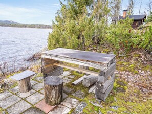 A rustic wooden bench with a metal top, surrounded by moss-covered stones and a backdrop of trees and a lake. - Rukaniemen kontio by Interhome (Kuusamo)