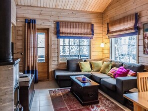 Ein gemütliches Wohnzimmer mit einer Couch, einem Couchtisch und einem Kamin. Es gibt Fenster mit Vorhängen, eine Tür und eine Holzbalkendecke.
