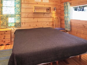 A wooden cabin room with a bed, a nightstand, a chair, and a window with curtains.