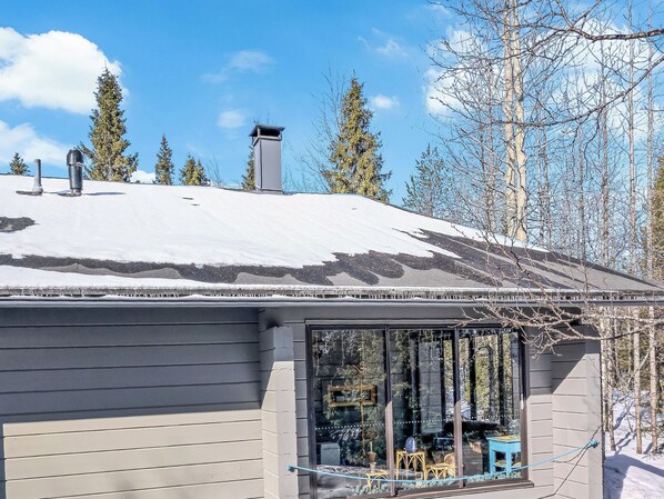 A house with a snow-covered roof, a chimney, and a window with a view of trees and a blue chair outside.