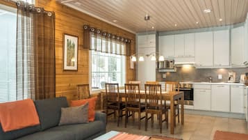 Un interior modern amb sostre de fusta, una zona de menjador amb cadires de fusta, una cuina amb armaris blancs i un sofà gris amb coixins taronja.