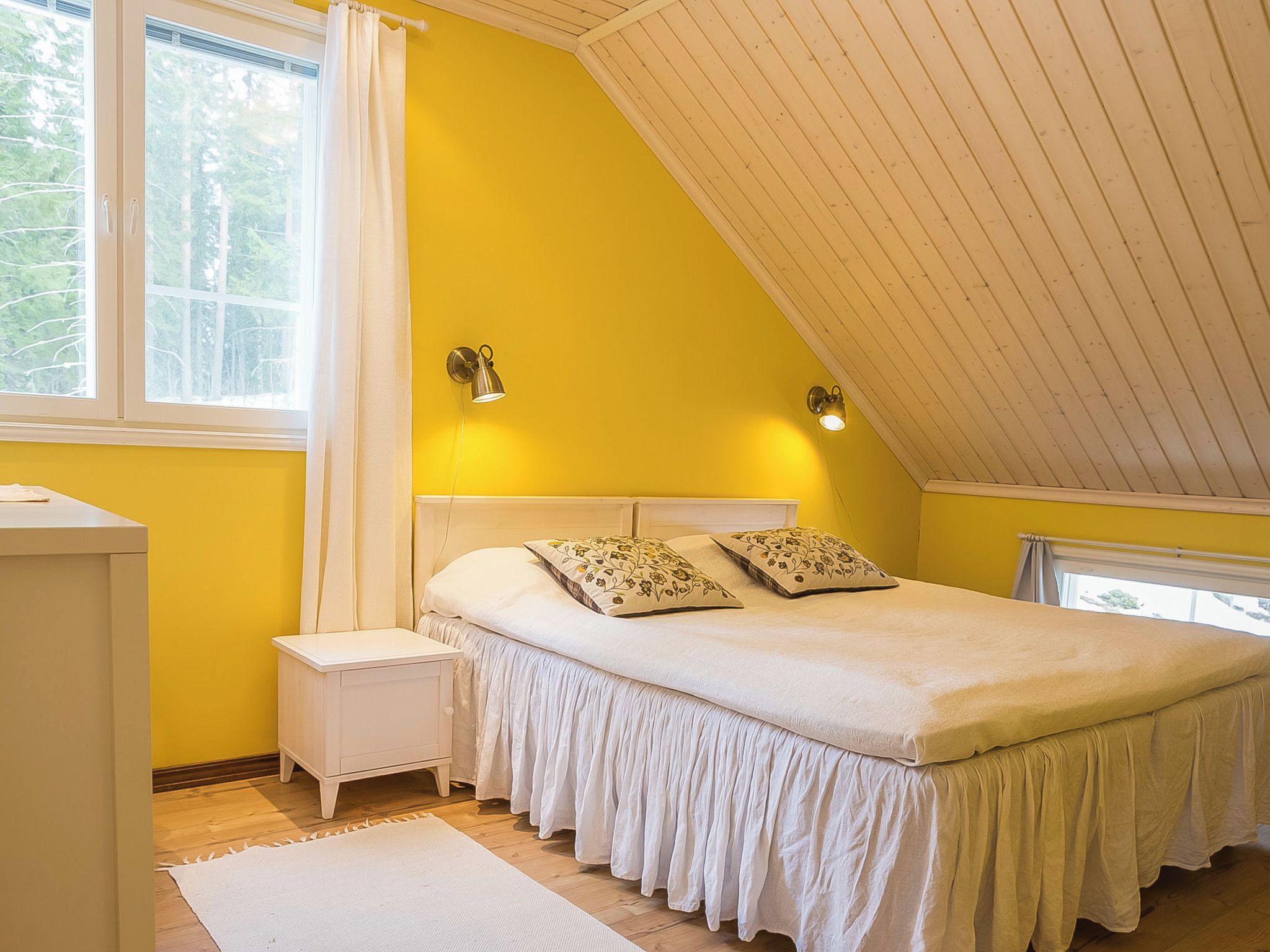 Une chambre à coucher avec un plafond en pente, un lit agrémenté d’oreillers à motifs floraux, une table de chevet et une fenêtre avec des rideaux.