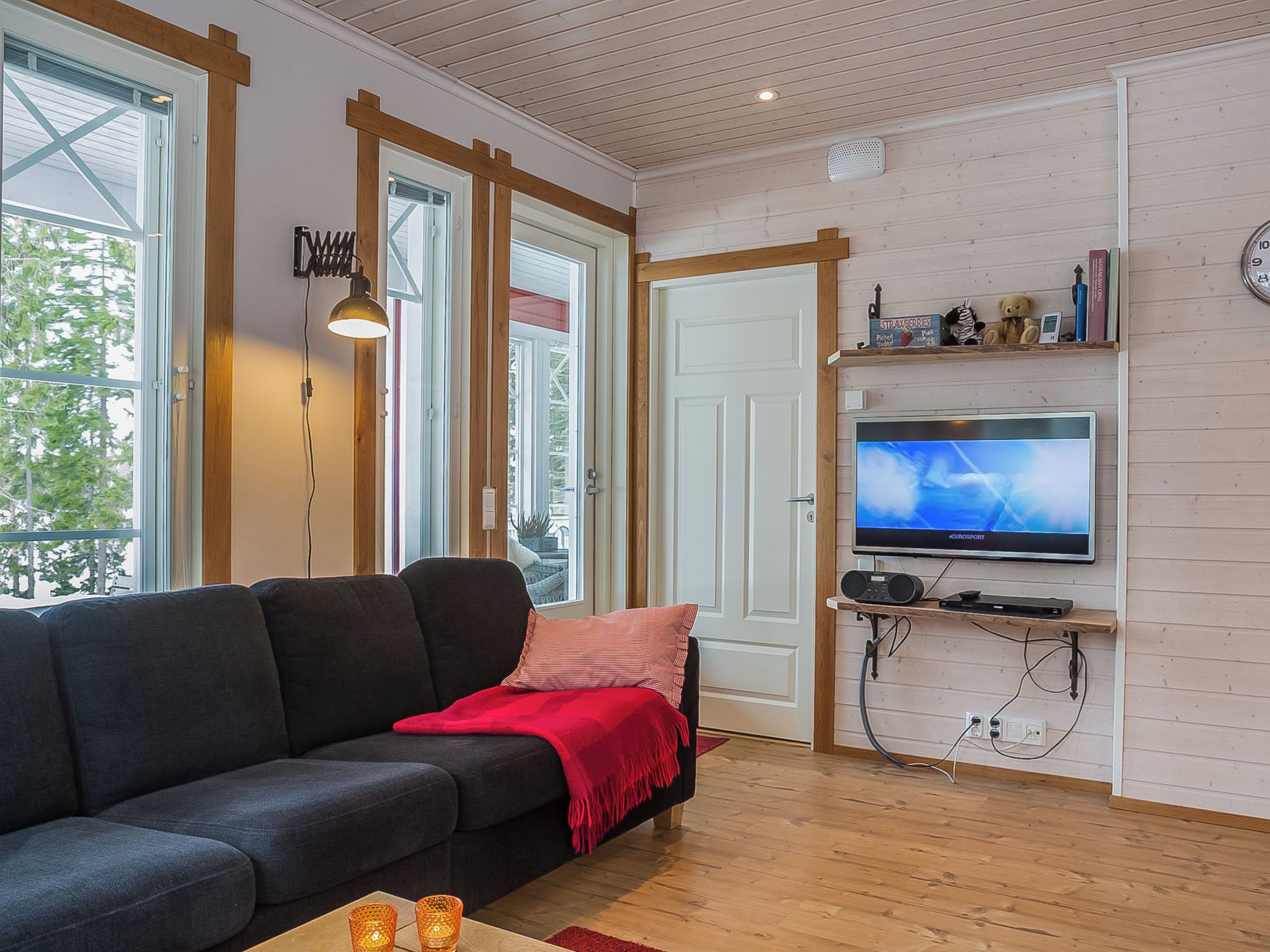 A modern living room with a dark sofa, a red throw blanket, a television on a wooden stand, and a large window with a view of trees.
