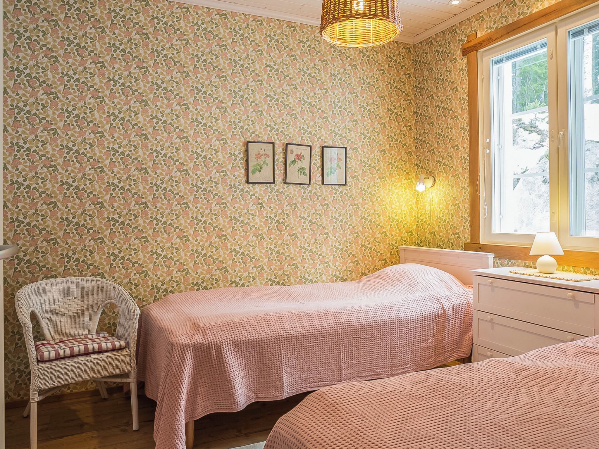 Ein Schlafzimmer mit floralem Wandpapier, zwei Betten, einem Korbsessel, einem Nachttisch mit Lampe und einem Fenster mit Blick.