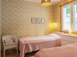 Un dormitorio con papel tapiz floral, dos camas, una silla de mimbre, una mesita de noche con una lámpara y una ventana con vistas.