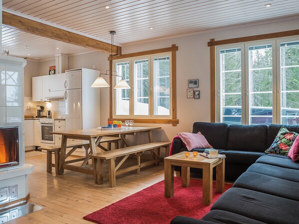 A cozy living room with a wooden table, a sofa, and a red rug. There is a kitchen area with cabinets and a refrigerator. - Villa kukkula by Interhome (Multia)