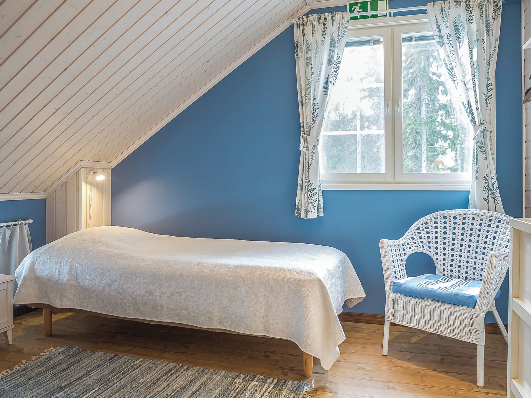 A bedroom with a bed, a wicker chair, a window with curtains, and a visible exit sign.