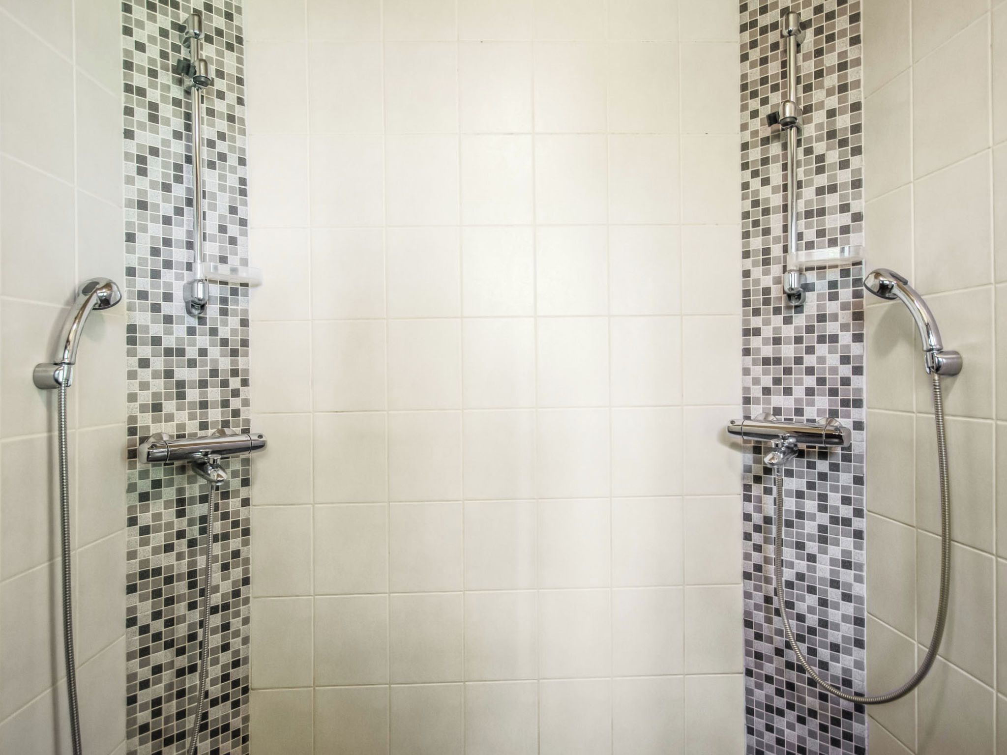 Ein Duschbereich mit zwei Chrom-Duschköpfen und Armaturen, vor einer mit Mosaikmuster versehenen Wand.
