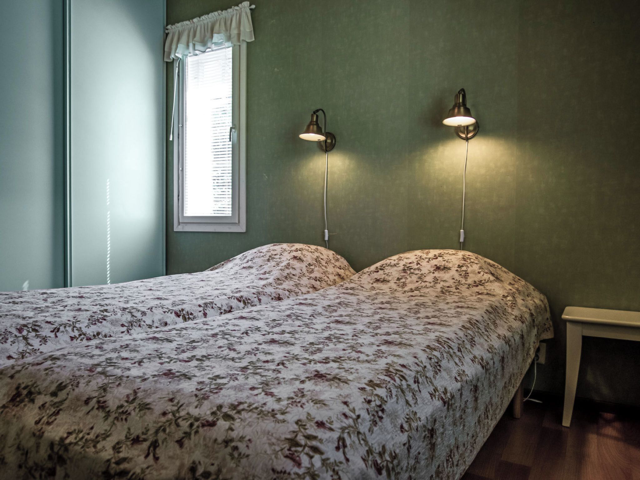 A bedroom with a bed, two wall-mounted lamps, a window with blinds, and a bedside table.
