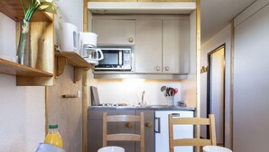Uma cozinha compacta com micro-ondas, pia e um balcão pequeno. Há duas cadeiras de madeira, uma mesa posta com pratos brancos, xícaras e uma cesta de croissants. Uma tigela de frutas está no lado esquerdo da mesa.