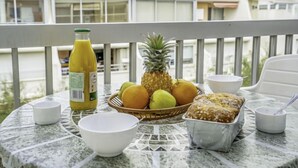Une table avec un panier de fruits, une miche de pain, une bouteille de jus et des bols sur un balcon.