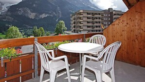 A balcony with a round table and two chairs, overlooking a mountain and a building with flags. - Chalet Abendrot apARTments by Interhome (Grindelwald)