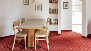 A dining area with a wooden table and chairs, a red patterned carpet, a bookshelf with books, and a kitchen visible through an open doorway. - Chalet Abendrot apARTments by Interhome (Grindelwald)