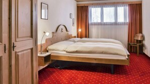 A bedroom with a wooden bed, a nightstand, a lamp, a window with curtains, and a red carpet. - Chalet Abendrot apARTments by Interhome (Grindelwald)
