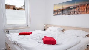 Un lit bien fait, avec du linge de lit blanc et rouge, une lampe de chevet et une œuvre d’art accrochée au mur représentant une scène de plage.
