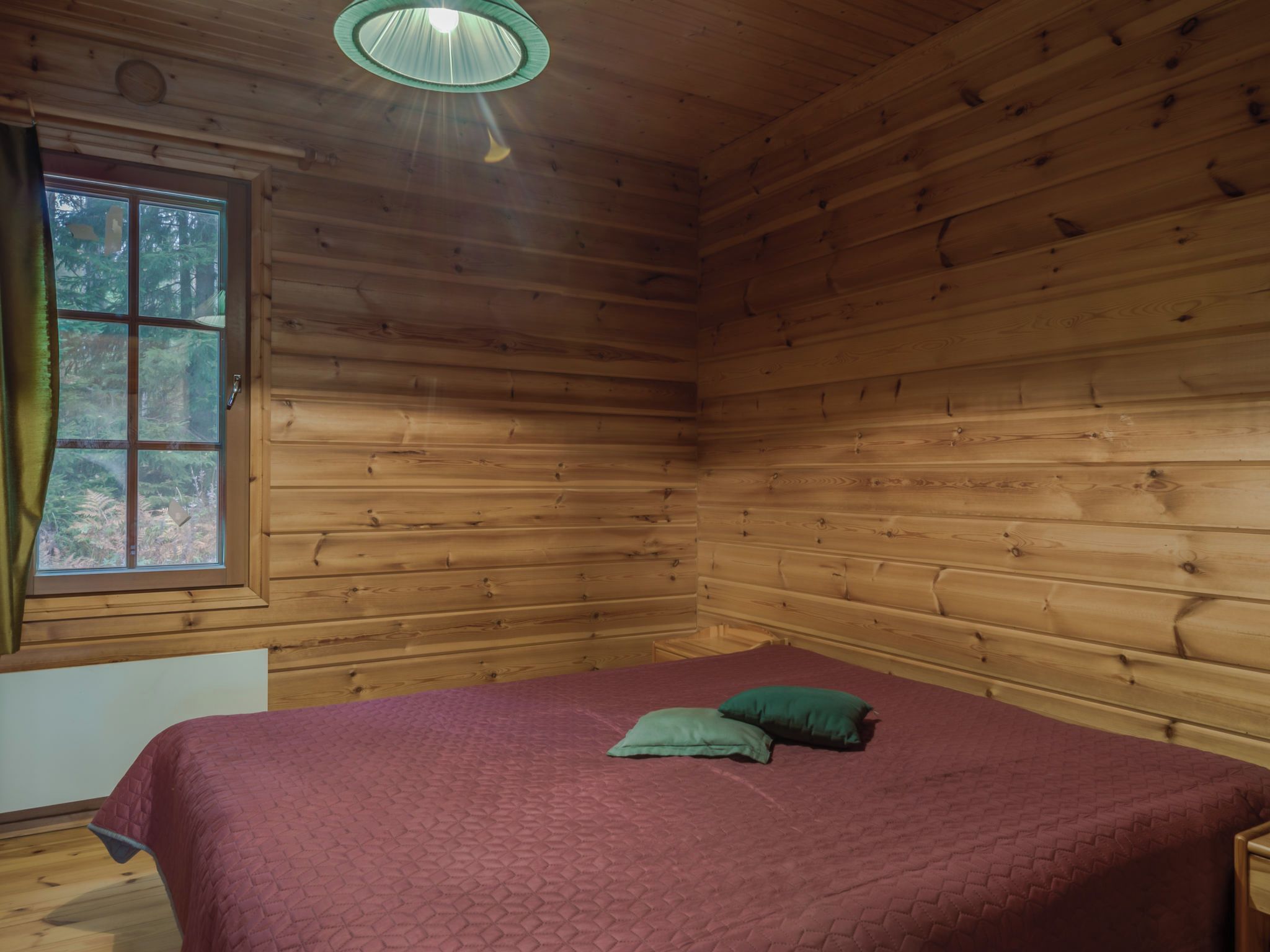 Una habitación con paredes de madera, una cama con colcha roja, una ventana con cortinas y una lámpara colgante.