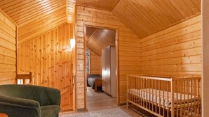 Una cabaña de madera con techo alto, una silla verde, una cuna y un baño a la vista al fondo.
