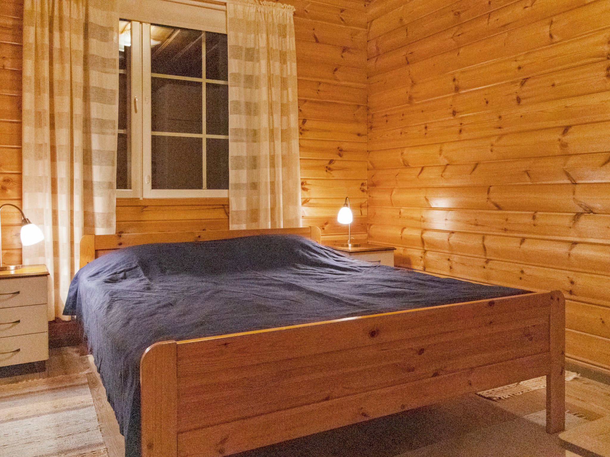 Un dormitorio de cabaña de madera con cama, mesitas de noche y una ventana con cortinas.