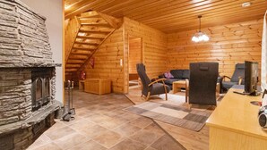 Interior de una cabaña de troncos con chimenea de piedra, techo de madera y una zona de estar acogedora con sofá y sillones.