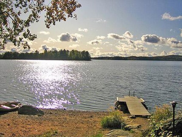 Näkymä järvelle, jossa puinen laituri ulottuu tyynen veden ylle. Laituria ympäröivät puut ja taivaalla on kirkasta.