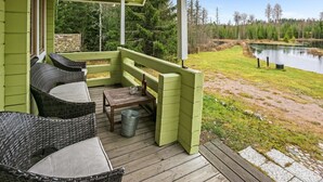A green wooden deck with wicker furniture, a small table, and a metal bucket. - Metsämökki by Interhome (Raasepori)