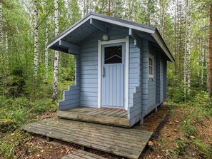 A small, single-story wooden structure with a front door and a small window, surrounded by a forest.