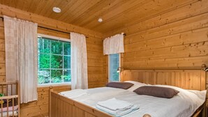 A wooden cabin bedroom with a bed, two pillows, a nightstand, a window with curtains, and a view of greenery outside. - Vacation home Kuutti by Interhome (Punkaharju)